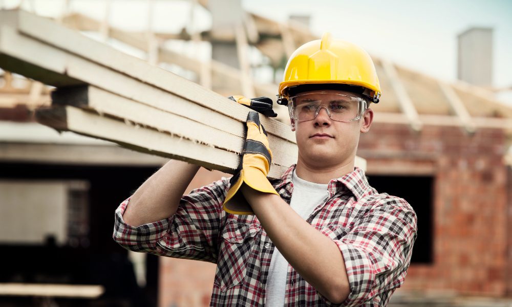 Человек в желтой строительной каске и с деревянными брусками на плече на строительстве дома