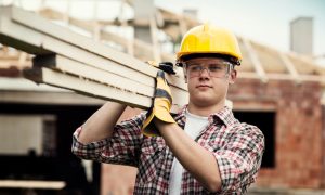 Человек в желтой строительной каске и с деревянными брусками на плече на строительстве дома