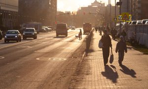 Яркое весенне солнце освещает улицу города, по дороге едут автомобили, по тротуару идут пешеходы.