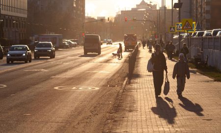 Яркое весенне солнце освещает улицу города, по дороге едут автомобили, по тротуару идут пешеходы.