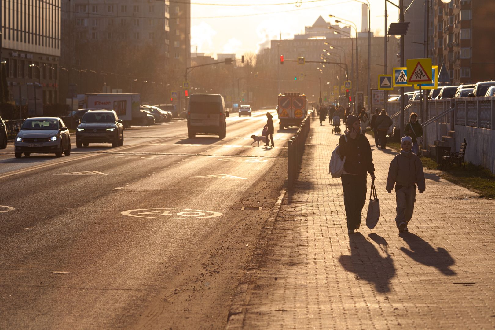 Яркое весенне солнце освещает улицу города, по дороге едут автомобили, по тротуару идут пешеходы.