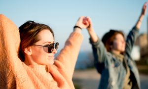 Две девушки подняли руки вверх, подставили лица солнцу и улыбаются.