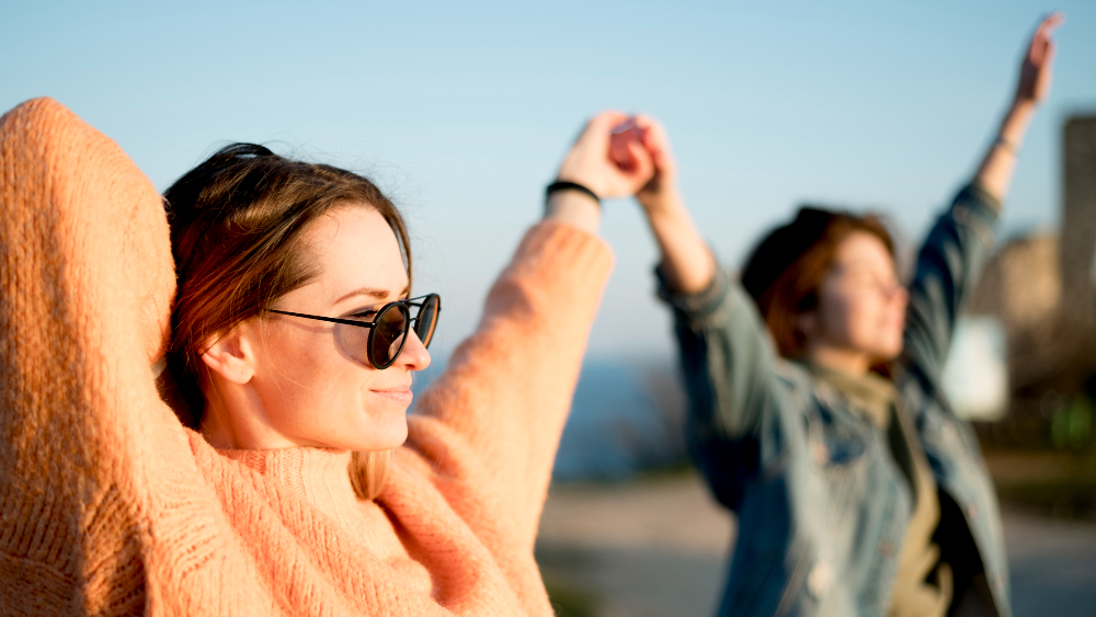 Две девушки подняли руки вверх, подставили лица солнцу и улыбаются.