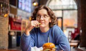 Девушка смотрит вдаль и задумчиво жует кусочек картофеля, не обращая внимания на бургер перед собой.