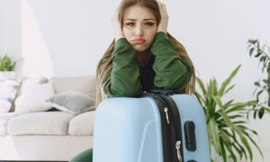 Грустная девушка в зеленом свитере рядом с чемоданом.