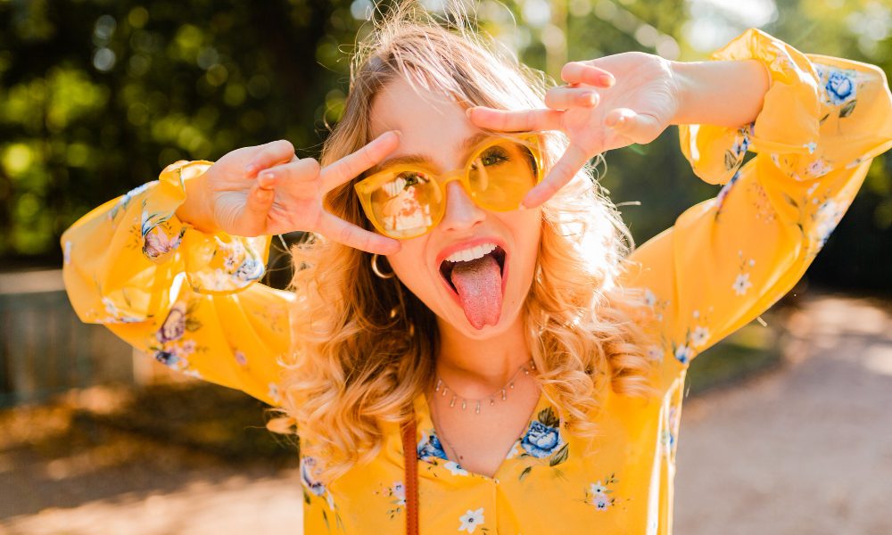 Девушка в желтых очках и блузке показывает язык и строит рожи.