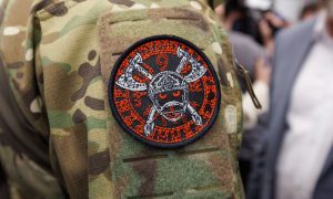 На изображении нашивка на камуфляжной одежде. На нашивке череп с мечами и красными буквами по кругу.