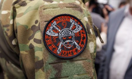 На изображении нашивка на камуфляжной одежде. На нашивке череп с мечами и красными буквами по кругу.