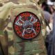 На изображении нашивка на камуфляжной одежде. На нашивке череп с мечами и красными буквами по кругу.